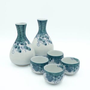 May include: A white ceramic sake set with a blue floral design. The set includes two bottles and four small cups. The bottles have a bulbous shape with a narrow neck. The cups are cylindrical. The design features blue grape-like clusters.