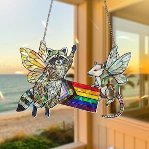 Könnte beinhalten: Dekorative Fensteraufhängung mit einem Waschbären und einem Opossum, beide mit Schmetterlingsflügeln. Der Waschbär winkt, und das Opossum hält eine Regenbogenflagge. Das Design besteht aus farbenfrohem, glasähnlichem Material.