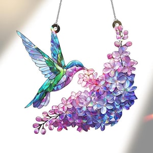 Puede incluir: Colibrí de vidriera con las alas extendidas, alimentándose de un racimo de flores moradas y rosas. El ave presenta tonos azules, verdes y morados. La obra de arte cuelga de dos cadenas plateadas.