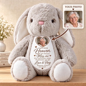 Op de afbeelding: Een grijze pluche konijnknuffel met een wit hartvormig embleem op de borst. Het embleem toont een ronde foto van een oudere vrouw en de tekst: A HUG FROM HEAVEN WHEN YOU REALLY MISS ME HUG THIS BUNNY TIGHT I'M SENDING YOU MY LOVE & HUG MORNING, NOON AND NIGHT.