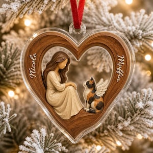 May include: A heart-shaped ornament with a wood-grain finish featuring a sculpted figure of a person with long brown hair in a cream dress sitting beside a calico cat with white angel wings. The names Nicole and Muffin are etched on the sides.