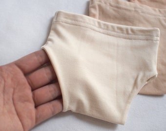 May include: Two pairs of beige cotton underwear, one pair is folded and the other is held in a hand.
