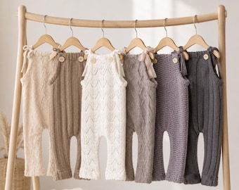 May include: A collection of six knitted rompers hanging on wooden hangers. The rompers feature various textures, including cable knit and openwork patterns, in shades of beige, cream, taupe, and gray. Each piece has shoulder ties and buttons.