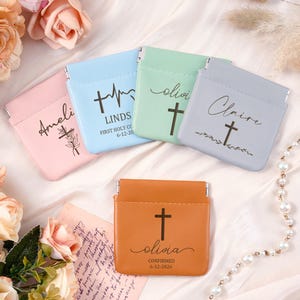 May include: A collection of personalized coin purses in pastel colors: pink, blue, green, and gray. Each purse features a cross design and a name, with additional text on some. An orange purse with a cross and the name "Olivia" is also visible.