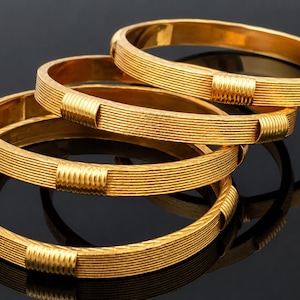 Könnte beinhalten: Vier goldfarbene Armreifen mit geripptem Design und rechteckigen Akzenten. Die Armreifen sind gestapelt und spiegeln sich auf einer dunklen Oberfläche wider, was ihre polierte Oberfläche und die aufwendigen Details zeigt. Klassischer Schmuck.