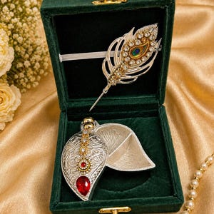 May include: A silver-toned feather-shaped brooch and a leaf-shaped pendant with a red gemstone are displayed inside a dark green velvet box. The brooch is adorned with clear and emerald-colored crystals. The pendant has a red gemstone and gold accents.