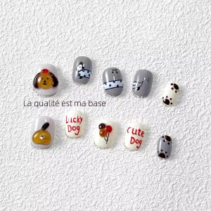 May include: A set of ten artificial nails with dog-themed designs. The nails are painted in white, grey, and brown, with illustrations of dogs and the words "Lucky Dog" and "Cute Dog". The French text "La qualité est ma base" is also visible on the image.