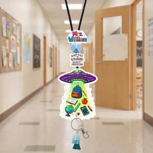 Könnte beinhalten: Ein Schlüsselband mit einem lila UFO-Motiv, das Schulmaterialien wie einen Rucksack, ein Buch, einen Apfel, einen Globus, ein Lineal, einen Taschenrechner und einen Stift zeigt. Es enthält ein Schild mit Mrs. William, Don't Talk To Strangers Safety Education, eine blaue Perle und eine Quaste.