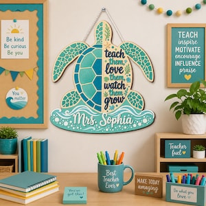 Op de afbeelding: Een decoratieve houten wandhanger in de vorm van een zeeschildpad, uitgevoerd in teal en blauwe kleuren met de tekst 'teach them, love them, watch them grow' en 'Mrs. Sophia' op de voorzijde, opgehangen aan een wit touw.