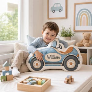 May include: A plush throw pillow shaped like a vintage blue race car with a tan seat, black wheels, and the name William printed in black script on the side. The pillow features a detailed illustration of a car with a steering wheel and fenders.