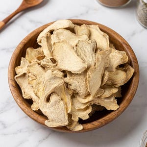 Può includere: Una ciotola di legno piena di fette di zenzero essiccato. Lo zenzero è di colore marrone chiaro e ha una consistenza leggermente rugosa. La ciotola è fatta di legno marrone chiaro e poggia su una superficie di marmo bianco. Un cucchiaio di legno è visibile sullo sfondo.
