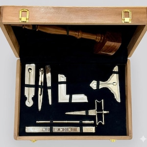 Puede incluir: Una caja de madera con interior de terciopelo negro, que contiene un mazo de madera y varias herramientas masónicas de color plateado. Las herramientas incluyen una escuadra, un compás y una regla. La caja tiene cierres dorados.