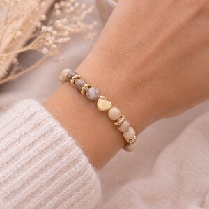 Könnte beinhalten: Ein Perlenarmband bestehend aus runden, marmorierten beigen und braunen Steinperlen, die mit kleinen, polierten goldfarbenen Metallzwischenperlen und einem zentralen, glatten, herzförmigen Anhänger in Goldoptik kombiniert sind.