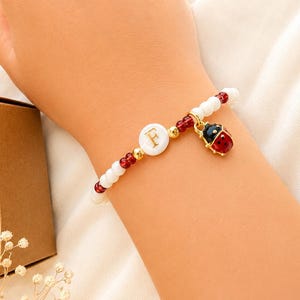 Peut inclure: Un bracelet de perles composé de perles blanches et rouges translucides, une perle centrale blanche circulaire avec la lettre F dorée, et une petite breloque coccinelle dorée avec des détails en émail rouge et noir, porté au poignet.