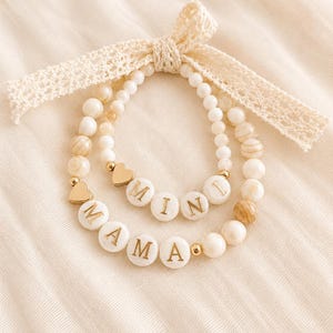 Peut inclure: Deux bracelets de perles composés de perles blanches et beiges marbrées, de breloques en forme de cœur dorées et de perles rondes blanches avec des lettres dorées. Un bracelet porte l'inscription MAMA et l'autre MINI. Un ruban de dentelle crème est noué.
