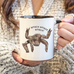Könnte beinhalten: Weiße Keramik-Tasse mit schwarzem Rand und Henkel. Die Tasse zeigt eine Cartoon-Illustration einer braunen Kreatur mit dem Text "amaze amaze amaze!" in Gelb.