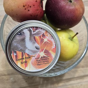 May include: A glass bowl holds several apples of varying colors, including red, yellow, and a speckled pink. A jar of "Cayenne Apple Butter" sits in the bowl, with a goat illustration on the lid.