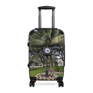 Peut inclure: Une valise à roulettes arborant une impression photographique d'une horloge de rue noire située dans un parc. L'image montre un grand palmier, de l'herbe verte et un parterre de fleurs blanches au pied du mât. La valise possède une poignée télescopique.