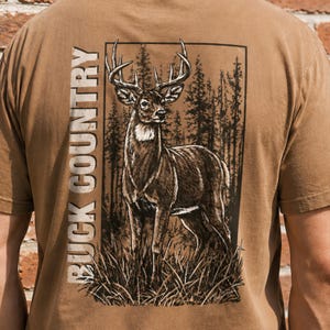 May include: A tan t-shirt featuring a rectangular graphic on the back. The graphic depicts a deer standing in a forest with tall grass in the foreground. The text BUCK COUNTRY is printed vertically in a bold, white, sans-serif font to the left.