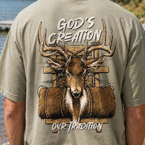 May include: A sage green t-shirt featuring a graphic on the back of a deer head with large antlers positioned in front of a wooden structure and a rolled brown fur blanket. The text reads GOD'S CREATION and OUR TRADITION in white lettering.