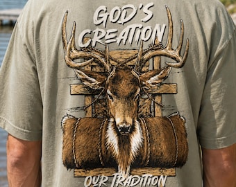 May include: A sage green t-shirt featuring a graphic on the back of a deer head with large antlers positioned in front of a wooden structure and a rolled brown fur blanket. The text reads GOD'S CREATION and OUR TRADITION in white lettering.