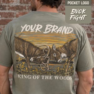 May include: An olive green t-shirt featuring a graphic on the back of two deer locking antlers. The text reads "YOUR BRAND" at the top and "KING OF THE WOODS" at the bottom. A small inset shows the text "POCKET LOGO" and "BUCK FIGHT".