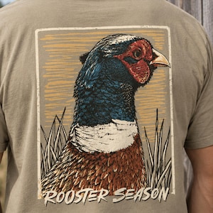 Może przedstawiać: Beżowy t-shirt z prostokątną grafiką na plecach. Nadruk przedstawia szczegółową ilustrację głowy bażanta z niebieskimi, czerwonymi i brązowymi piórami, na złotym tle z trawą oraz napisem ROOSTER SEASON.
