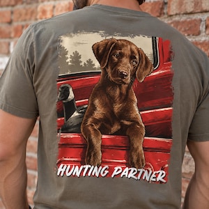 May include: An olive green t-shirt featuring a graphic on the back of a brown Labrador puppy and a Canada goose sitting in the bed of a red pickup truck. The text "HUNTING PARTNER" is printed in white capital letters at the bottom of the graphic.