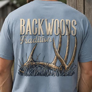 Op de afbeelding: Een lichtblauw t-shirt met een grafische print op de achterkant. Het ontwerp bevat de tekst "BACKWOODS" in grote, dikke letters boven het woord "Tradition" in een cursief lettertype. Onder de tekst staat een illustratie van een gewei in het gras.