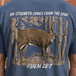 Pode incluir: Uma t-shirt azul com uma impressão gráfica de um veado castanho numa floresta. O texto "MY STRENGTH COMES FROM THE LORD" está impresso a branco acima do veado, e "PSALM 28:7" está impresso a branco abaixo da ilustração do veado.