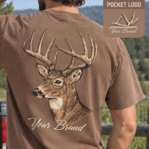 Può includere: Una t-shirt marrone con una grande grafica di una testa di cervo con corna sul retro e il testo Your Brand. L'area del petto anteriore include un logo da taschino più piccolo con corna di cervo e il testo POCKET LOGO e Your Brand.