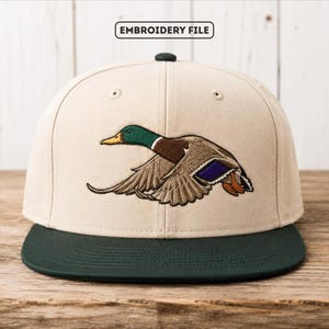 May include: A beige baseball cap with a dark green flat brim and an embroidered mallard duck in flight on the front panel. The duck features a green head, brown body, and purple wing patch. The text EMBROIDERY FILE is centered above the cap.