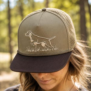 Puede incluir: Una gorra tipo trucker de color verde oliva con visera plana marrón oscuro y un detalle de cuerda en la base de la corona. El panel frontal muestra un bordado blanco de un perro de muestra en la hierba, con paneles traseros de malla.