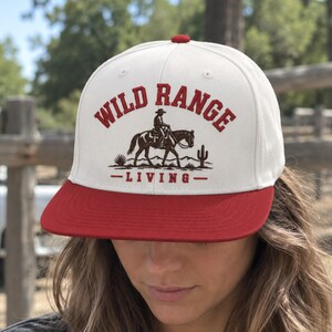 Peut inclure: Une casquette snapback blanche avec une visière rouge et un bouton rouge sur le dessus. L'avant présente un motif brodé d'une personne à cheval dans un paysage désertique avec un cactus, accompagné du texte rouge WILD RANGE LIVING.