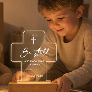 May include: A cross-shaped acrylic night light with a wooden base, featuring the engraved text "Be Still AND KNOW THAT I AM GOD PSALM 46:10" and a small cross icon at the top. The light emits a warm glow, illuminating the etched white lettering.