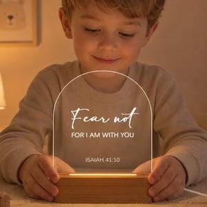 Può includere: Una luce notturna in acrilico ad arco con base in legno, caratterizzata dal testo stampato in bianco Fear not FOR I AM WITH YOU ISAIAH 41:10. La luce emette un bagliore caldo dalla base, illuminando il pannello in acrilico trasparente.