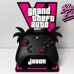 Op de afbeelding: Een zwarte gaming controller standaard met een groot roze V-logo met de tekst Grand Theft Auto. De standaard bevat zwarte palmbomsilhouetten, een voet met roze accenten en de naam Jason in witte letters op de voorzijde van de controller.