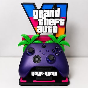 Può includere: Un supporto per controller di videogiochi con un controller wireless viola poggiato su una base con la scritta 'YOUR-NAME'. Il supporto mostra il logo di Grand Theft Auto VI in blu, rosa e arancione, accentuato da due sagome di palme verde brillante.