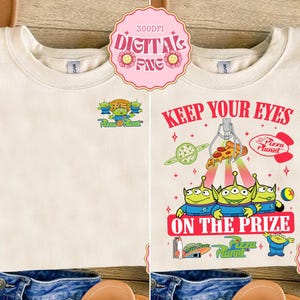 Könnte beinhalten: Cremefarbenes Sweatshirt mit Grafiken aus dem Animationsfilm Toy Story. Eine Seite zeigt das Pizza Planet-Logo und Aliens, die andere Seite den Text "KEEP YOUR EYES ON THE PRIZE" mit Alien- und Pizzastück-Illustrationen.