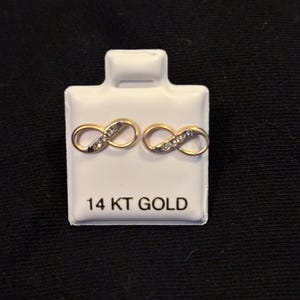 May include: A pair of gold-colored infinity symbol stud earrings with small, clear stones. The earrings are displayed on a white card that reads "14 KT GOLD" in black text. The background is a dark, solid color.