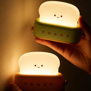 May include: Two novelty toast-shaped night lights. Each light has a glowing, white bread slice with a smiling face, set in a green and orange toaster base. The word "TOAST" is printed on the front of each base.
