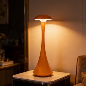 Peut inclure: Lampe de table orange avec une tige longue et élancée et un dessus arrondi. La lampe est posée sur une table de chevet à dessus en marbre avec une base noire et une bordure dorée. La lampe émet une lueur chaleureuse.