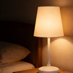 May include: A white bedside lamp with a beige lampshade is illuminated, casting a warm glow. The lamp sits on a wooden nightstand next to a bed with a white comforter. The room is dimly lit.