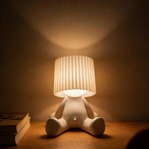 May include: A white, whimsical lamp shaped like a seated figure with a pleated lampshade. The lamp sits on a wooden surface, casting a warm glow. A stack of books is visible to the left. The lamp's design is modern and playful.