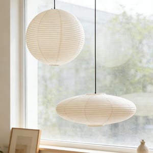 Puede incluir: Dos lámparas colgantes con pantallas de papel blanco cuelgan del techo. Una es una esfera, la otra es ovalada. Las pantallas tienen líneas horizontales. Las luces cuelgan frente a una ventana.