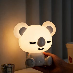 Op de afbeelding: Een koala-vormig nachtlampje met een zacht, gloeiend wit lichaam en grijze accenten. De lamp heeft gesloten ogen en een aan/uit-knop. De oren zijn een lichtere tint wit. De lamp wordt door een hand vastgehouden.