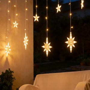 May include: String lights with warm white star-shaped lights hanging from a beam. The lights are strung in vertical lines, with some stars having a multi-pointed design. A white couch is in the background.