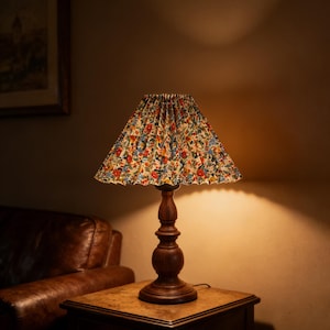 Könnte beinhalten: Tischlampe mit floralem Lampenschirm. Die Lampe hat einen dunklen Holzfuß und steht auf einem kleinen Holztisch. Der Lampenschirm zeigt ein farbenfrohes Muster aus roten, blauen und gelben Blumen. Der Tisch ist etwa 60 cm hoch.