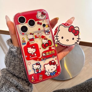 May include: A red and clear phone case featuring Hello Kitty designs and a matching pop socket. The case has a red border and showcases various Hello Kitty illustrations, including the character on a bus and holding a strawberry. The pop socket has a white background with Hello Kitty's face.