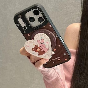 May include: A black phone case with a brown backing, decorated with small white stars. A heart-shaped pop socket features a bear and bunny illustration. The phone case is held by a person.